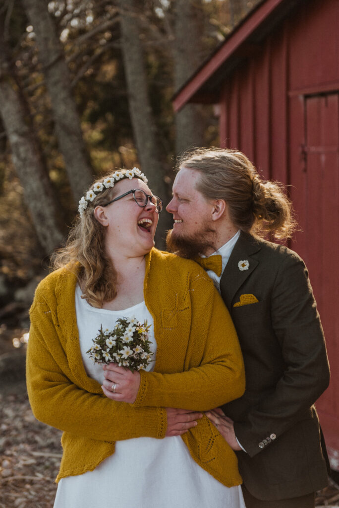 Brudpar skrattar hjärtligt framför röd byggnad. Bruden har egenstickad kofta och virkade blommor i håret.
